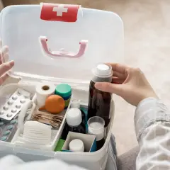 Otwarta apteczka pierwszej pomocy z lekami, plastrami i bandażami. Dłoń sięga po buteleczkę z płynem.